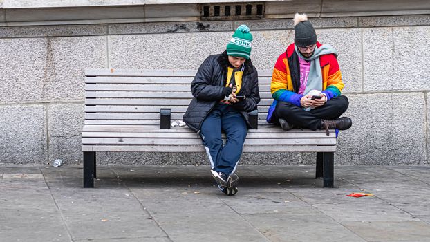 London, UK - January 1, 2020: Two men sit on a bench on the side of the street and look at their smartphones