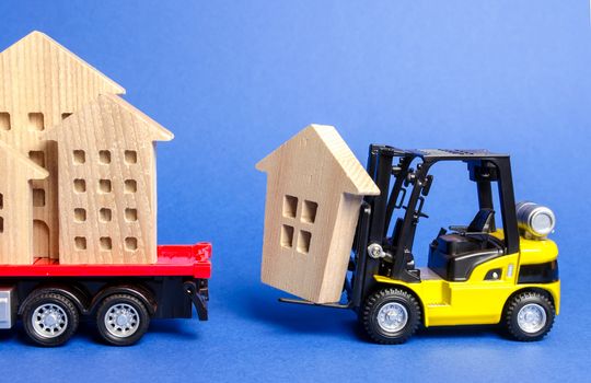 A yellow forklift loads a wooden figure of a house into a truck. Concept of transportation and cargo shipping, moving company. Construction of new houses and objects. Industry. Move entire buildings.