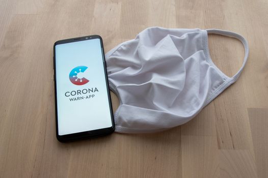 Zittau, Saxony / Germany - June 5th 2020: Smartphone with splash screen of German Corona Warn contact tracing App with white face mask on wooden surface.