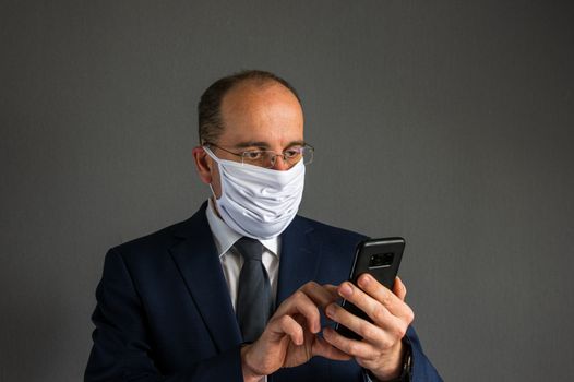 portrait of a young business man wearing a suit and tie and protective face mask checks information on his smartphone in front of gray background. Lots of copy space