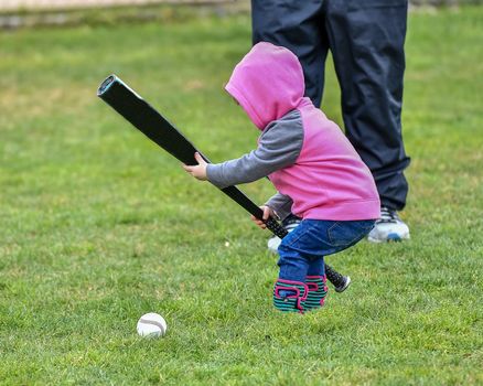 Adorable infant playing with a baseball bat and ball at the baseball park. Cute baby with headband trying to pick up the baseball and also trying to hit the ball with a bat that is too big for her.