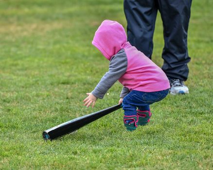 Adorable infant playing with a baseball bat and ball at the baseball park. Cute baby with headband trying to pick up the baseball and also trying to hit the ball with a bat that is too big for her.