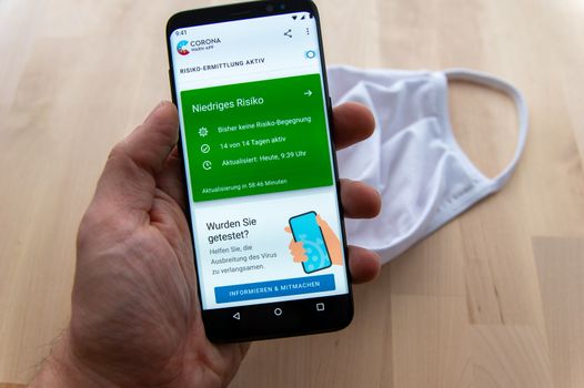 Zittau, Saxony / Germany - June 5th 2020: Hand holding Smartphone showing active Risk analysis of German Corona Warn App with blurred white face mask in background. Niedriges Risiko = Low Risk