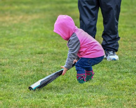 Adorable infant playing with a baseball bat and ball at the baseball park. Cute baby with headband trying to pick up the baseball and also trying to hit the ball with a bat that is too big for her.