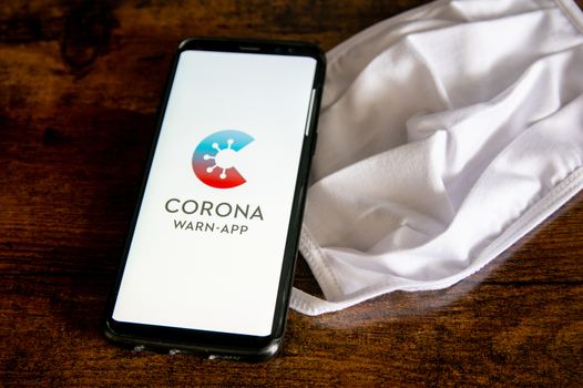 Zittau, Saxony / Germany - June 4th 2020: German Corona Virus Warn App for COVID-19 tracing with white face mask on wooden surface. The new normal, items for everyday life in 2020