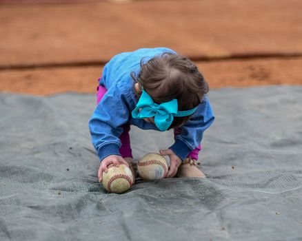 Adorable infant playing with a baseball bat and ball at the baseball park. Cute baby with headband trying to pick up the baseball and also trying to hit the ball with a bat that is too big for her.