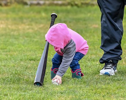 Adorable infant playing with a baseball bat and ball at the baseball park. Cute baby with headband trying to pick up the baseball and also trying to hit the ball with a bat that is too big for her.