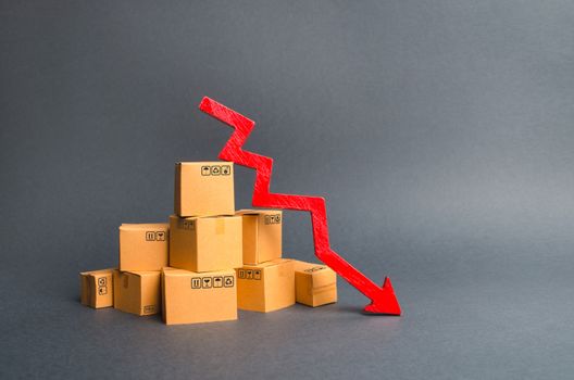 A pile of cardboard boxes and a red arrow down. The decline in the production of goods and products, the economic downturn and recession. Falling consumer demand, declining exports or imports.