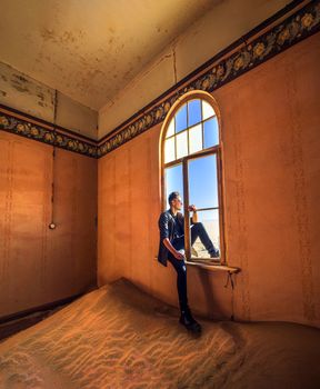 Young tourist sitting in the window of an empty room located in the ruins of the ghost town Kolmanskop in the Namib desert near Luderitz, Namibia, Southern Africa