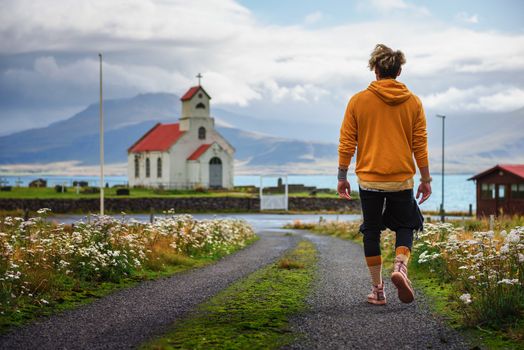 Young man walking on a dirt road towards a church and a cemetery in Iceland with a lake in the background. Loneliness, hope, sadness , depression and solitude concept