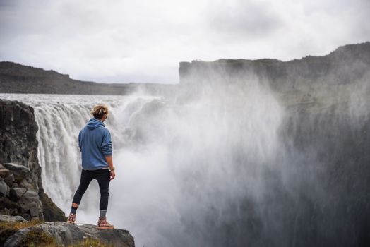 Young hiker standing at the edge of the Dettifoss waterfall located on the Jokulsa a Fjollum river in Iceland. Dettifoss is the second most powerful waterfall in Europe after the Rhine Falls.