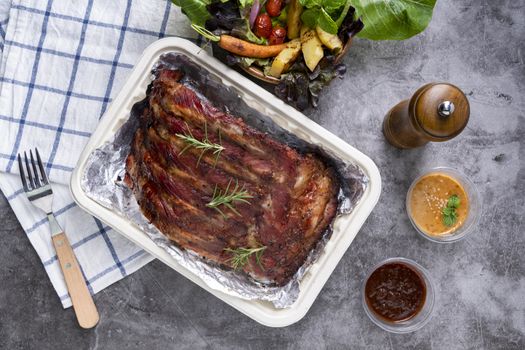 Closeup of pork ribs grilled with BBQ sauce and caramelized in honey with Vegetables and sauces on a drak table, prepared food.