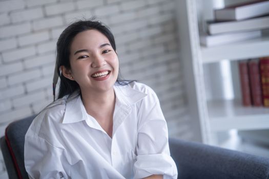 beautiful young asian woman smiling while sitting on the chair against white brick wall