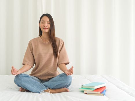 Young woman meditating on bed at home after reviewing the textbook. Morning fitness, mindfulness concept.