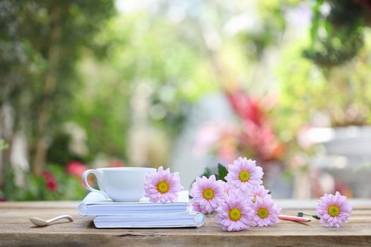 White coffee cup with white notebook and pink pencil with pink Chrysanthemum flower on wooden table at outdoor