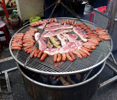 A street vendor cooks sausages and slabs of pork on a large barbecue rack
