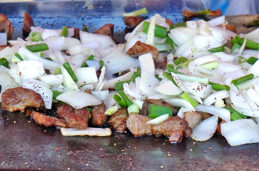 An outdoor vedor cooks meat, onions and leeks on a Teppanyaki griddle
