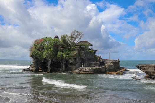 Waves shatter on a cliff at the top of which is the Hindu temple of Tanah Lot. Temple built on a rock in the sea off the coast of Bali island, Indonesia. Lush tropical vegetation on top of a cliff.