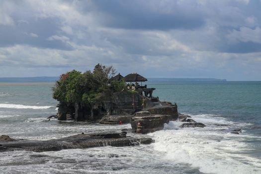 Aerial view of Tanah Lot temple on cliff in Indian ocean. Waves are breaking on a rock cliff off the coast of Bali. Hindu temple built on a rock in the sea. Lush tropical vegetation on top of a cliff.