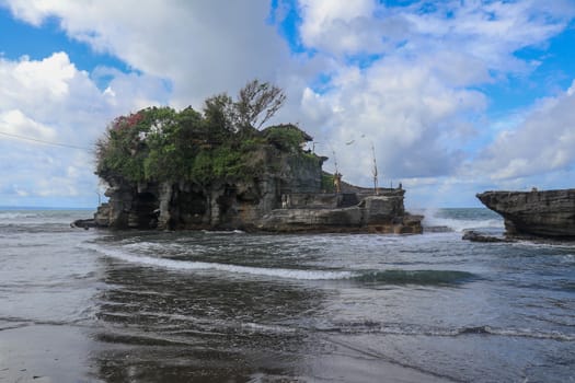 Waves shatter on a cliff at the top of which is the Hindu temple of Tanah Lot. Temple built on a rock in the sea off the coast of Bali island, Indonesia. Lush tropical vegetation on top of a cliff.