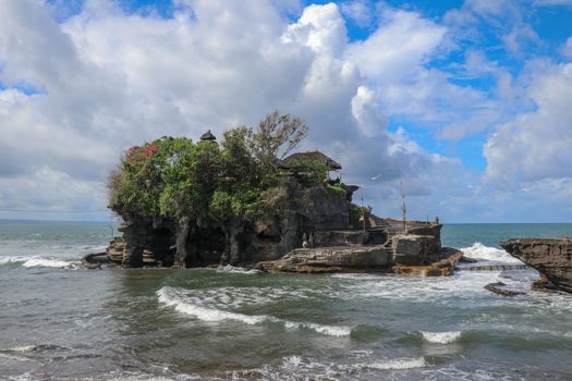 Waves shatter on a cliff at the top of which is the Hindu temple of Tanah Lot. Temple built on a rock in the sea off the coast of Bali island, Indonesia. Lush tropical vegetation on top of a cliff.