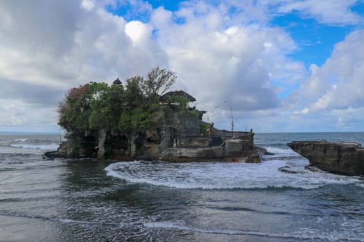 Waves shatter on a cliff at the top of which is the Hindu temple of Tanah Lot. Temple built on a rock in the sea off the coast of Bali island, Indonesia. Lush tropical vegetation on top of a cliff.