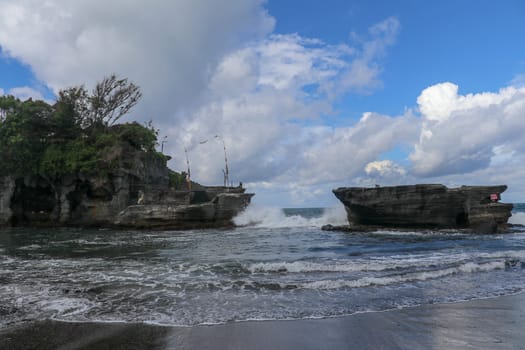 Waves shatter on a cliff at the top of which is the Hindu temple of Tanah Lot. Temple built on a rock in the sea off the coast of Bali island, Indonesia. Lush tropical vegetation on top of a cliff.