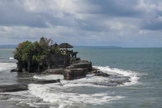 Waves shatter on a cliff at the top of which is the Hindu temple of Tanah Lot. Temple built on a rock in the sea off the coast of Bali island, Indonesia. Lush tropical vegetation on top of a cliff.