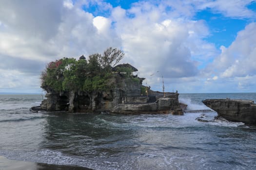 Waves shatter on a cliff at the top of which is the Hindu temple of Tanah Lot. Temple built on a rock in the sea off the coast of Bali island, Indonesia. Lush tropical vegetation on top of a cliff.