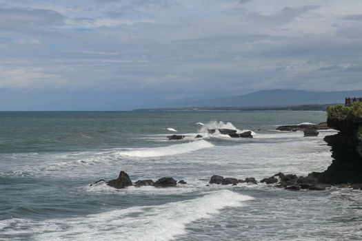 Rock cliffs in the Indian Ocean off the coast of Bali island, near Batu Bolong and tourism attraction spot Tanah Lot temple. Waves are shattering on rocks of solidified black volcanic lava. Indonesia.