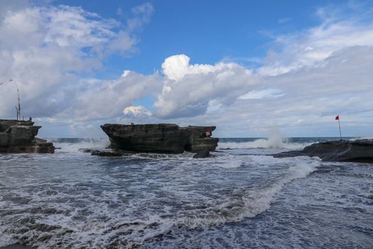Sea surf and waves hit the rock cliffs at Tanah Lot Temple. Sea water turns into white foam. Red warning flag highlights dangers of ocean. Unbridled ocean energy. Natural background for your project.