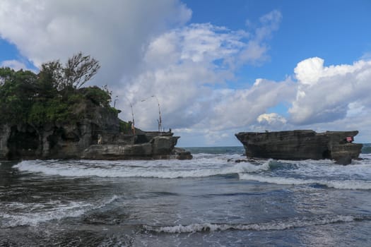 Waves shatter on a cliff at the top of which is the Hindu temple of Tanah Lot. Temple built on a rock in the sea off the coast of Bali island, Indonesia. Lush tropical vegetation on top of a cliff.