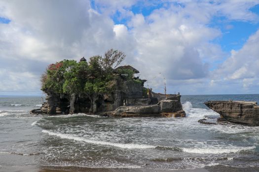 Waves shatter on a cliff at the top of which is the Hindu temple of Tanah Lot. Temple built on a rock in the sea off the coast of Bali island, Indonesia. Lush tropical vegetation on top of a cliff.