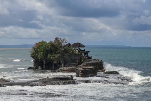 Aerial view of Tanah Lot temple on cliff in Indian ocean. Waves are breaking on a rock cliff off the coast of Bali. Hindu temple built on a rock in the sea. Lush tropical vegetation on top of a cliff.