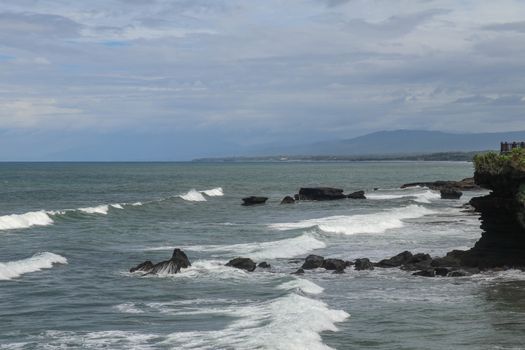 Rock cliffs in the Indian Ocean off the coast of Bali island, near Batu Bolong and tourism attraction spot Tanah Lot temple. Waves are shattering on rocks of solidified black volcanic lava. Indonesia.