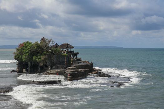 Aerial view of Tanah Lot temple on cliff in Indian ocean. Waves are breaking on a rock cliff off the coast of Bali. Hindu temple built on a rock in the sea. Lush tropical vegetation on top of a cliff.