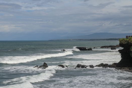 Rock cliffs in the Indian Ocean off the coast of Bali island, near Batu Bolong and tourism attraction spot Tanah Lot temple. Waves are shattering on rocks of solidified black volcanic lava. Indonesia.