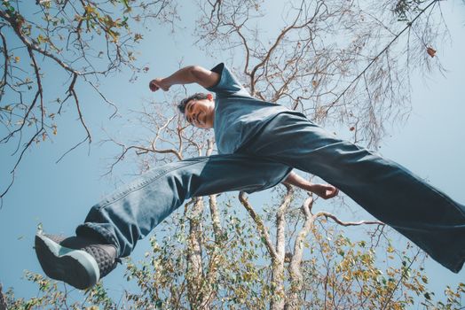 Low angle view of man midair by jumping, crossing step over the camera shot below in forest with tree and sky overhead in concept travel, active lifestyle, overcome obstacles in life or future