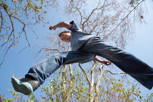 Low angle view of man midair by jumping, crossing step over the camera shot below in forest with tree and sky overhead in concept travel, active lifestyle, overcome obstacles in life or future