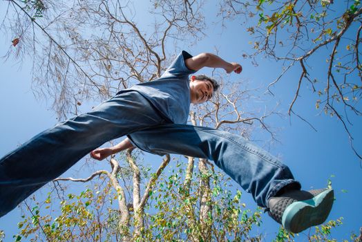 Low angle view of man midair by jumping, crossing step over the camera shot below in forest with tree and sky overhead in concept travel, active lifestyle, overcome obstacles in life or future