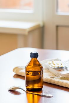 A cough syrup bottle with a spoon, a thermometer and some pills blister on the table close to the window
