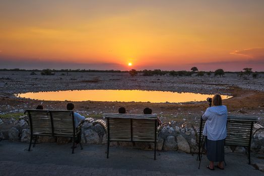 Okaukuejo, Namibia - April 3, 2019 : Tourists wait for wildlife at the waterhole of Okaukuejo Campsite in Etosha National Park, Namibia, with beautiful sunset in the background.