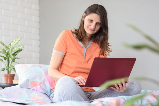 The girl in pajamas works in the computer sitting at home on the bed