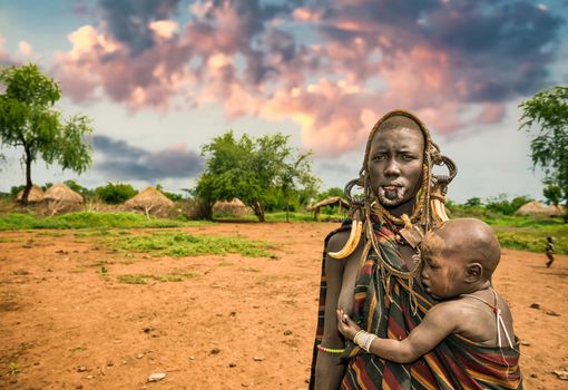 OMO VALLEY, ETHIOPIA - MAY 7, 2015 : Woman from the african tribe Mursi with her baby and traditional horns in Mago National Park, Ethiopia