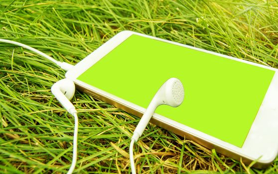 Headphones and a White phone on the green grass. Chromakey green screen of a mobile phone. Banner template with text space.