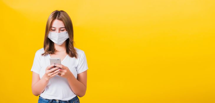 Portrait Asian Thai beautiful happy young woman wearing mask protection, reading news update on smartphone health care, studio shot isolated on yellow background, with copy space