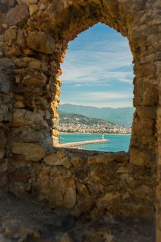 Window in the fortress in the stone wall. View of landscape and lghthouse of Alanya port. Alanya peninsula, Antalya district, Turkey, Asia