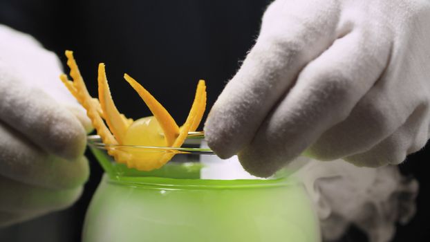 Cooking Grape Martini Cocktail. Close up bartenders hands decorating with orange zest. The glass is wrapped in steam