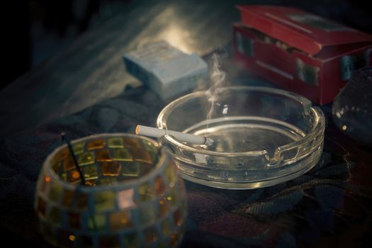 Smoke emitted from a burnt cigarette in a glass ashtray. It placed on a tablecloth on a wooden table. Close up, loop with selective focus.