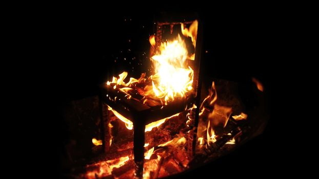Wooden chair burning inside the bonfire. Close up on sparks rises from the fire with heatwaves at night. A horror concept of a demonic ritual or devil and demons religious worship.